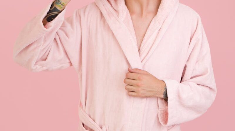 Young man in a pink bathrobe brushing teeth in front of a pink background.